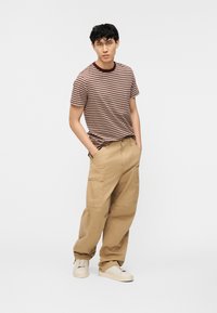 Νέος άνδρας που στέκεται με τα χέρια στις τσέπες, φοράει καφέ ριγέ μπλουζάκι, άνετο μπεζ παντελόνι cargo και μπεζ αθλητικά παπούτσια, σε λευκό φόντο.