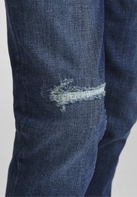 Jean en denim bleu foncé avec un patch usé bien visible au niveau du genou, présentant un bord effiloché et un accent en fil bleu clair.