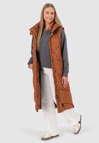 Gilet imbottito lungo di colore brown terra, con colletto alto, tasche con zip e un design leggero e trapuntato. Indossato sopra una felpa grigia.