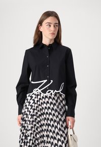 Camicia nera con bottoni e maniche lunghe, con un logo bianco sul petto, abbinata a una gonna plissettata nera e crema.