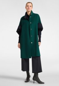 Abrigo verde de mezcla de lana con un diseño estructurado, que presenta un cuello alto, botones plateados y mangas anchas. Combinado con pantalones negros y botas de punta.
