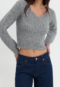 Pull gris en maille avec col en V et manches longues, présentant une texture côtelée. Associé à un jean taille haute bleu foncé.