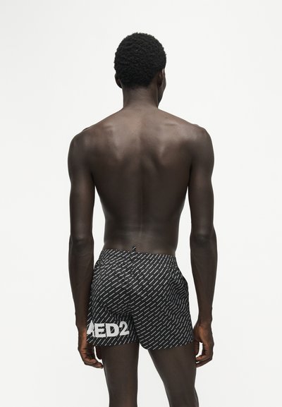 Homme torse nu à la peau foncée vu de dos portant un short noir à motifs avec le texte blanc "DSQUARED2" sur le côté gauche.