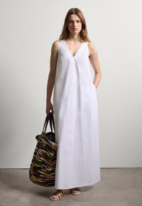 Weißes Leinen-Maxikleid mit V-Ausschnitt und Schlitzen, kombiniert mit einer mehrfarbigen geflochtenen Tragetasche. Schnüren-Sandalen runden den Look ab.