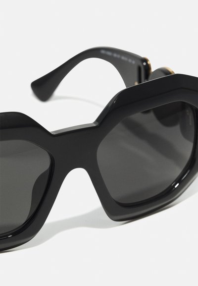 Svarta överdimensionerade solglasögon med geometrisk ram, blank yta, rökiga linser och böjda, eleganta bågar med guldaccent.
