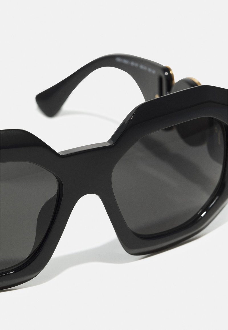 Occhiali da sole neri oversize con montatura geometrica, finitura lucida, lenti fumè e aste curve e affusolate con dettagli dorati.
