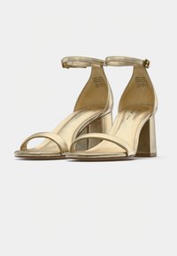 Χρυσά μεταλλικά σανδάλια με ψηλό τακούνι, ανοιχτό δάχτυλο, δύο μπροστινά λουριά, λουρί στον αστράγαλο και τετράγωνο τακούνι. Ομαλή υφή με ελάχιστες λεπτομέρειες.