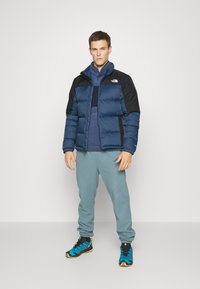 Giacca a vento blu navy con spalle nere, finitura testurizzata, con zip frontale, indossata sopra un pullover blu scuro, abbinata a pantaloni blu chiaro e sneakers blu vivaci.