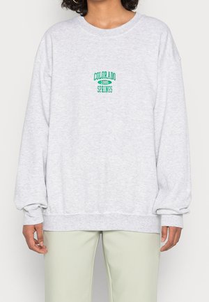 Persoon draagt een lichtgrijze trui met groene tekst "Colorado 1985 Springs", gecombineerd met lichtgekleurde broek.