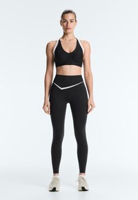 Jeune femme debout, portant un soutien-gorge de sport noir et blanc et des leggings, avec des baskets blanches, sur un fond clair uni.