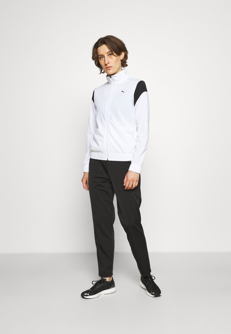 Weißer Zip-Up-Trainingsanzug-Oberteil mit schwarzen Schulterakzenten, kombiniert mit schwarzen tapered Pants und schwarzen Sportschuhen. Glatte Stofftextur.