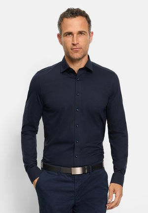 Man draagt een nauwsluitend donkerblauw overhemd met knopen, ingestopt in bijpassende broek met een zwarte riem, staand tegen een effen lichte achtergrond.