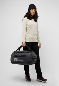Bolsa de deporte gris con detalles negros, fabricada con un tejido resistente, asas dobles y logo de "Jack Wolfskin". El modelo lleva una camiseta de manga larga crema y pantalones negros.