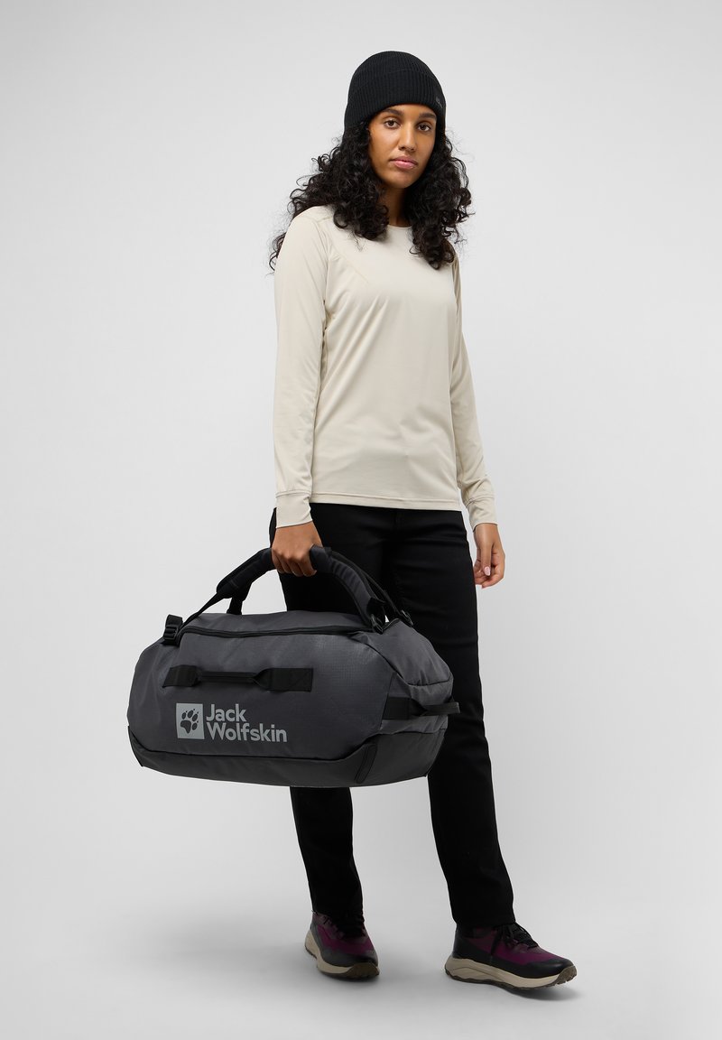 Bolsa de deporte gris con detalles negros, fabricada con un tejido resistente, asas dobles y logo de "Jack Wolfskin". El modelo lleva una camiseta de manga larga crema y pantalones negros.
