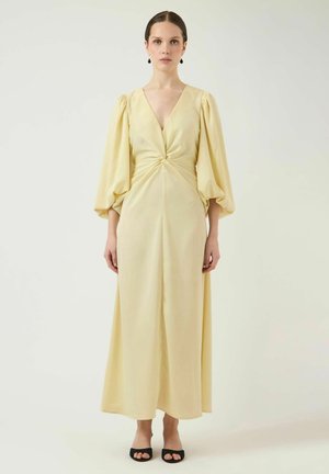 Kobieta w długiej, jasnopomarańczowej sukni z głębokim dekoltem w kształcie litery V, bufkami na długich rękawach, z detalem skręconej talii, w czarnych, otwartych na palce szpilkach, stojąca na jednolitym tle.