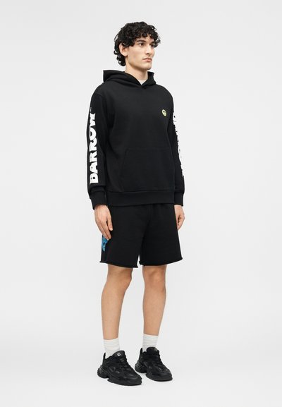 Ung mann med krøllete hår iført en svart hettegenser med hvit "BARROW"-tekst på ermene, svarte shorts, hvite sokker og svarte joggesko som står.