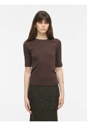 Joven mujer con cabello largo y oscuro que lleva una blusa marrón de manga corta y una falda verde oscuro con estampado de leopardo sobre un fondo liso.