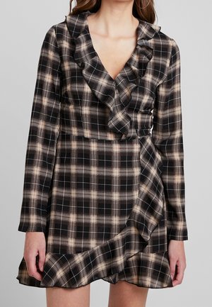 Frau trägt ein langärmeliges Kleid mit schwarzem und beigem Karomuster, einem gerafften V-Ausschnitt und einem asymmetrischen Volant-Saum.