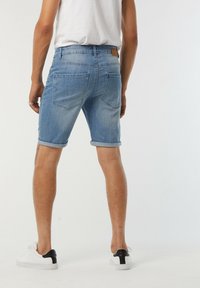 Shorts en denim bleu clair avec un ourlet roulé, comportant deux poches arrière et un patch en cuir marron sur la taille. Baskets blanches avec des accents noirs.