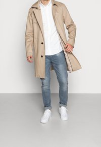 Trench beige con cierre de botón frontal, bolsillos laterales y cuello, llevado sobre una camisa blanca y jeans de un azul claro con zapatillas blancas.