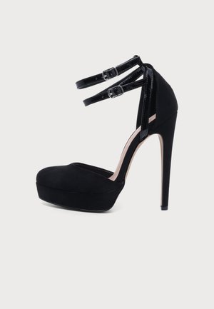 Chaussure noire à plateforme et à talon haut, avec un bout arrondi, un talon aiguille élevé et deux brides de cheville à boucles argentées.