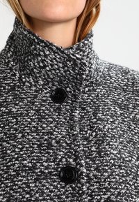 Tekstureret, grå og sort vævet jakke med en høj krave, der har to sorte knapper og et nærbillede af stoffets mønster.