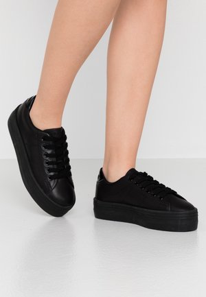 Beine in schwarzen niedrigen Plateau-Sneakern mit Schnürsenkeln, stehend auf einem glänzenden weißen Boden vor einem schlichten hellen Hintergrund.