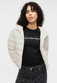 Ελαφρώς μπεζ μπουφάν πεφφέρ με φερμουάρ μπροστά και ψηλό γιακά, φορεμένο πάνω από ένα μαύρο t-shirt με λευκό κείμενο. Το σύνολο ολοκληρώνουν γκρι τζιν με σχέδιο.