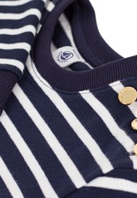 Streifenpullover in Marineblau und Weiß mit ribbelndem Ausschnitt und drei goldenen Knopfakzenten an der Schulter, gefertigt aus einem weichen Stoff.