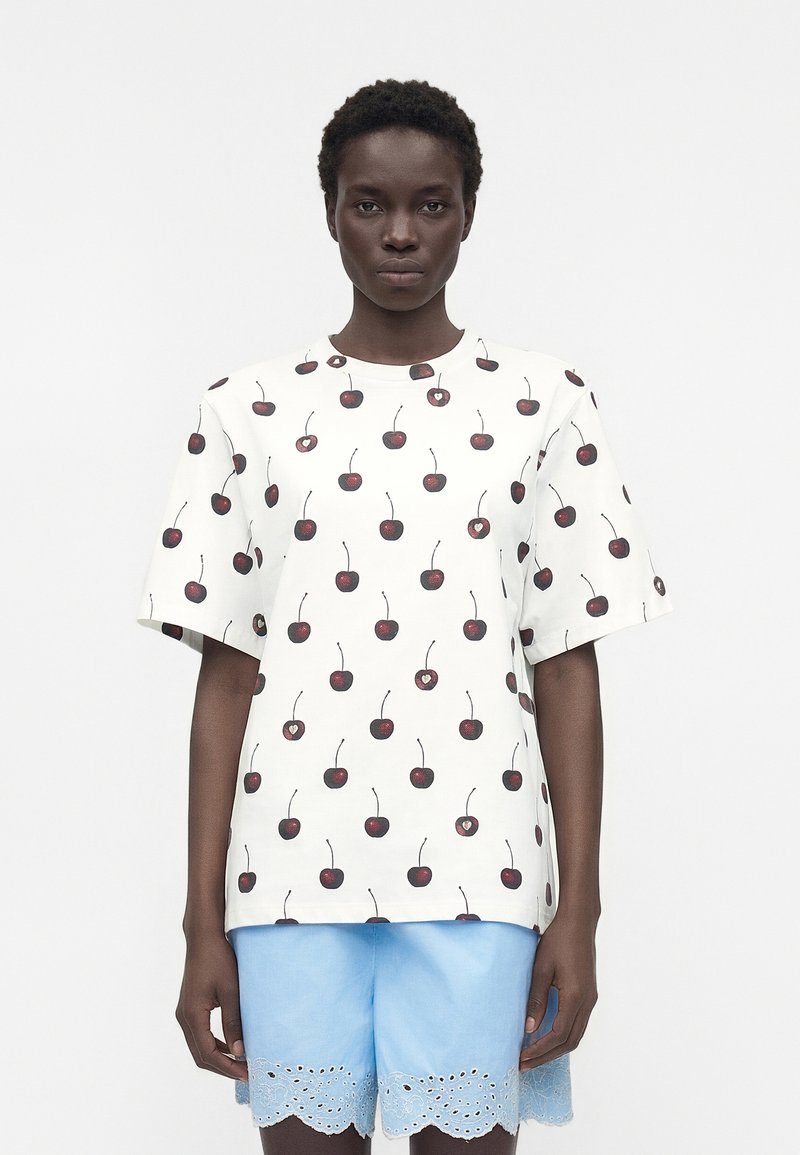 Άτομο με σκουρόχρωμο δέρμα που φοράει λευκό T-shirt με σχέδιο κερασιών και ανοιχτό μπλε σορτς με κυματιστό δαντελένιο τελείωμα, στέκεται μπροστά σε μονόχρωμο φόντο.