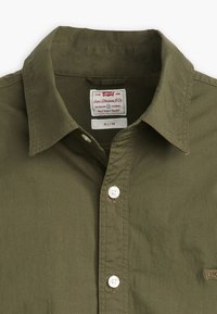 Olívzöld gombos ing, amely pamutból készült, slim fit szabású, hajtókás gallérral, fehér gombokkal, valamint egy címkével a márkáról és a stílusról.