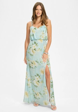 Kobieta stoi, ubrana w jasnoniebieską sukienkę maxi w kwiaty z cienkimi ramiączkami, bocznym rozcięciem oraz białe sandały na cienkich paskach na białym tle.