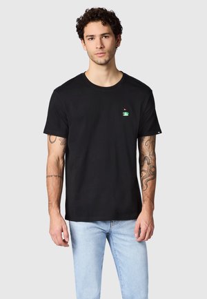 Man met krullend haar en tatoeages, die een zwart T-shirt draagt met een klein groen golfbaanlogo en lichtblauwe spijkerbroek, staand tegen een grijze achtergrond.