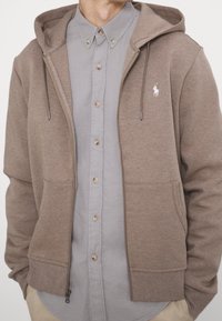 Felpa marrone con cappuccio, con zip frontale, tasche a marsupio e lacci, indossata sopra una camicia grigia chiara con bottoni.