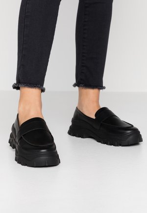 Personne portant des mocassins noirs épais et un jean noir à ourlet effiloché, debout sur un sol gris clair.