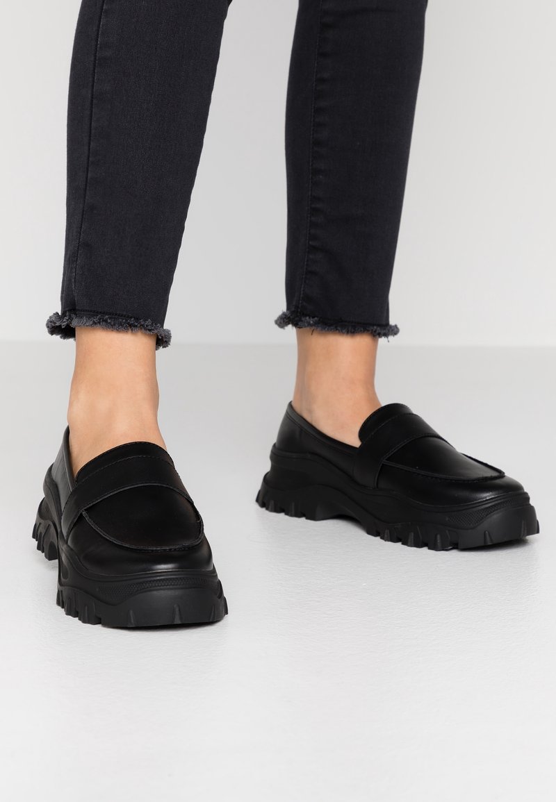 Personne portant des mocassins noirs épais et un jean noir à ourlet effiloché, debout sur un sol gris clair.