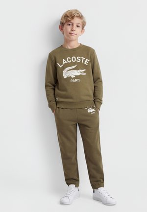 Lyshåret dreng står med hænderne i lommerne iført olivengrøn Lacoste sweatshirt og matchende bukser med hvide sneakers mod hvid baggrund.
