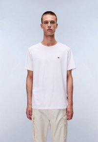 Valge puuvillane T-särk lühikeste varrukatega, ümmarguse kaelusega ja väike punane logo rinnal. Modell kannab seda helebeežide cargo-pükstega.