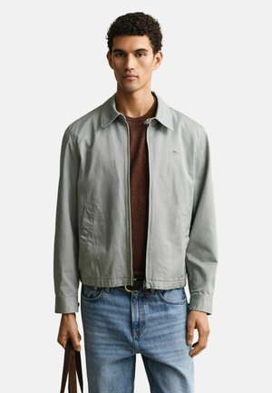 Uomo che indossa una giacca grigio chiaro con zip sopra una camicia marrone e jeans blu, che tiene la tracolla di una borsa marrone scuro con la mano sinistra.