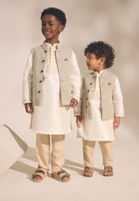 Deux enfants portent des tenues traditionnelles assorties comprenant des gilets beiges, des tuniques blanches avec des détails brodés, et des pantalons de couleur claire.