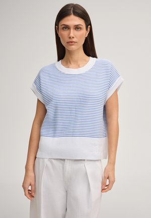 Frau mit glatten braunen Haaren trägt einen blau-weiß gestreiften Kurzarm-Pullover und weiße hoch taillierte Hosen, sie steht vor einem einfachen Hintergrund.