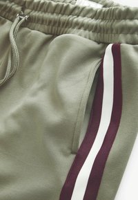 Olivgröna sweatpants med elastisk midja och dragband. Har sidofickor och vinröda och vita vertikala ränder längs sidorna.