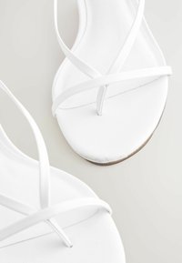 White strappy sandals with thin crossing straps and a toe post, shown in close-up on a light background.