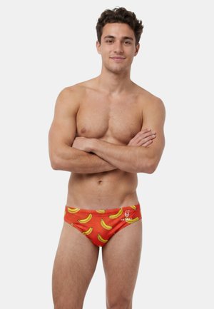 Jeune homme debout, les bras croisés, portant un slip de bain rouge orné de bananes jaunes sur un fond clair uni.