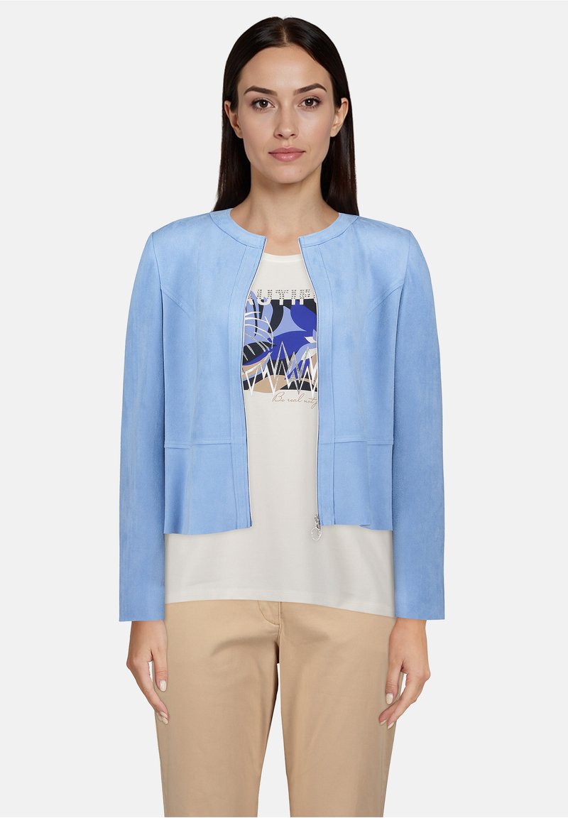 Veste en cuir bleu clair avec un col rond, des manches longues et une fermeture éclair à l'avant. Portée sur un t-shirt graphique blanc et un pantalon beige.