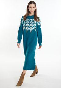 Türkisfarbenes Strickkleid mit einem weißen Blattmuster, ausgestattet mit langen Ärmeln und einer geraden Silhouette, kombiniert mit braunen Stiefeletten.