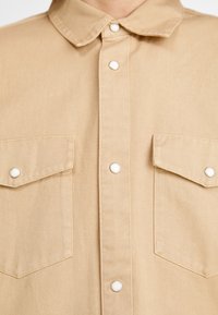 Beige paita, valmistettu kestävästä kankaasta, jossa on kauluksellinen pääntie, edessä painonapein suljettava etuosio ja kaksi rintataskua valkoisilla napeilla.