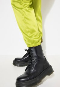 Czarne lace-up buty wojskowe z grubą podeszwą, zestawione z błyszczącymi, jasnobrączo zielonymi spodniami dresowymi z marszczonymi mankietami.