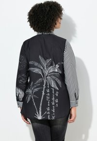 Sort skjorte med stribede ærmer; bagsiden har hvidt palmetrædesign og tekst. Bomuldsmateriale, afslappet pasform, med knapdetalje på manchetterne.