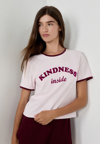 Giovane donna con lunghi capelli castani che indossa una maglietta bianca con bordi borgogna su cui è scritto "KINDNESS inside", in piedi contro un muro chiaro.